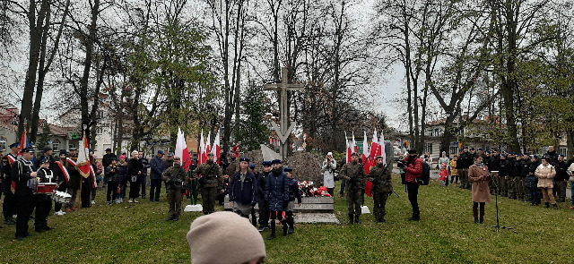 Pięcioro dzieci w mundurach harcerskich wracają od pomnika, przy którym  po obu stronach stoją  żołnierze, po lewej stronie stoi orkiestra dęta, po prawej stronie kobieta prowadząca uroczystość. W tle zebrany przy pomniku tłum. 