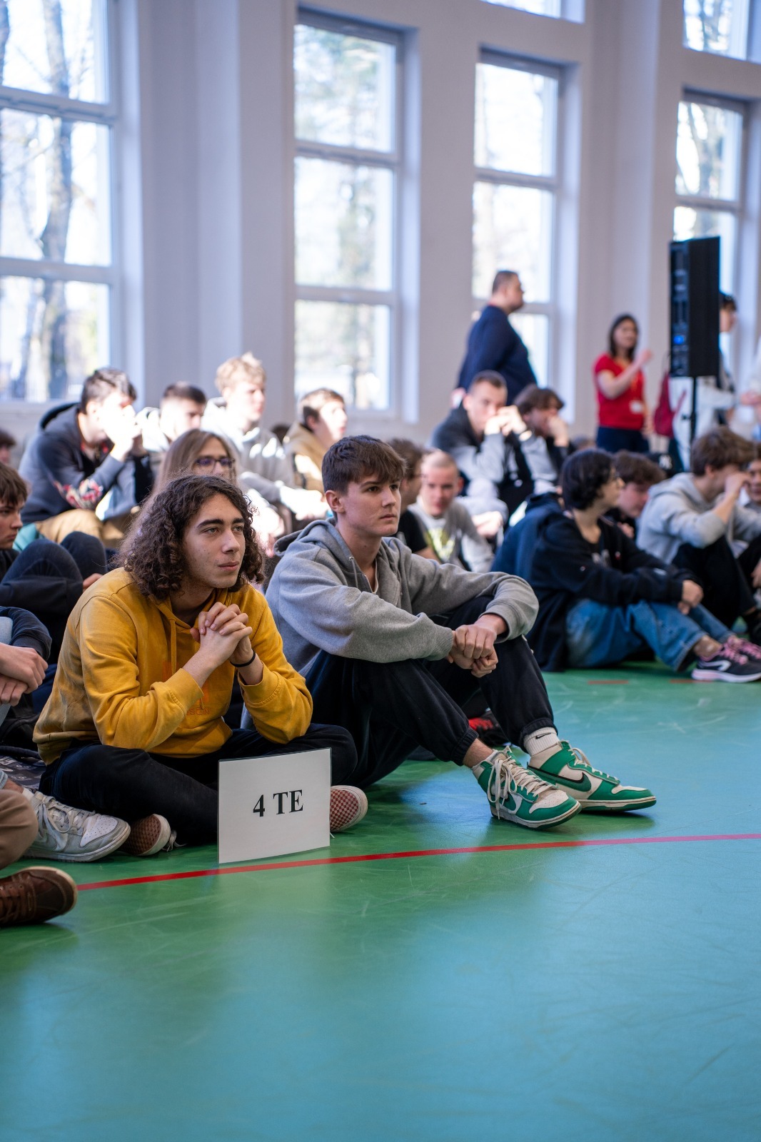 Uczniowie siedzący klasami na sali gimnastycznej w trakcie apelu. 