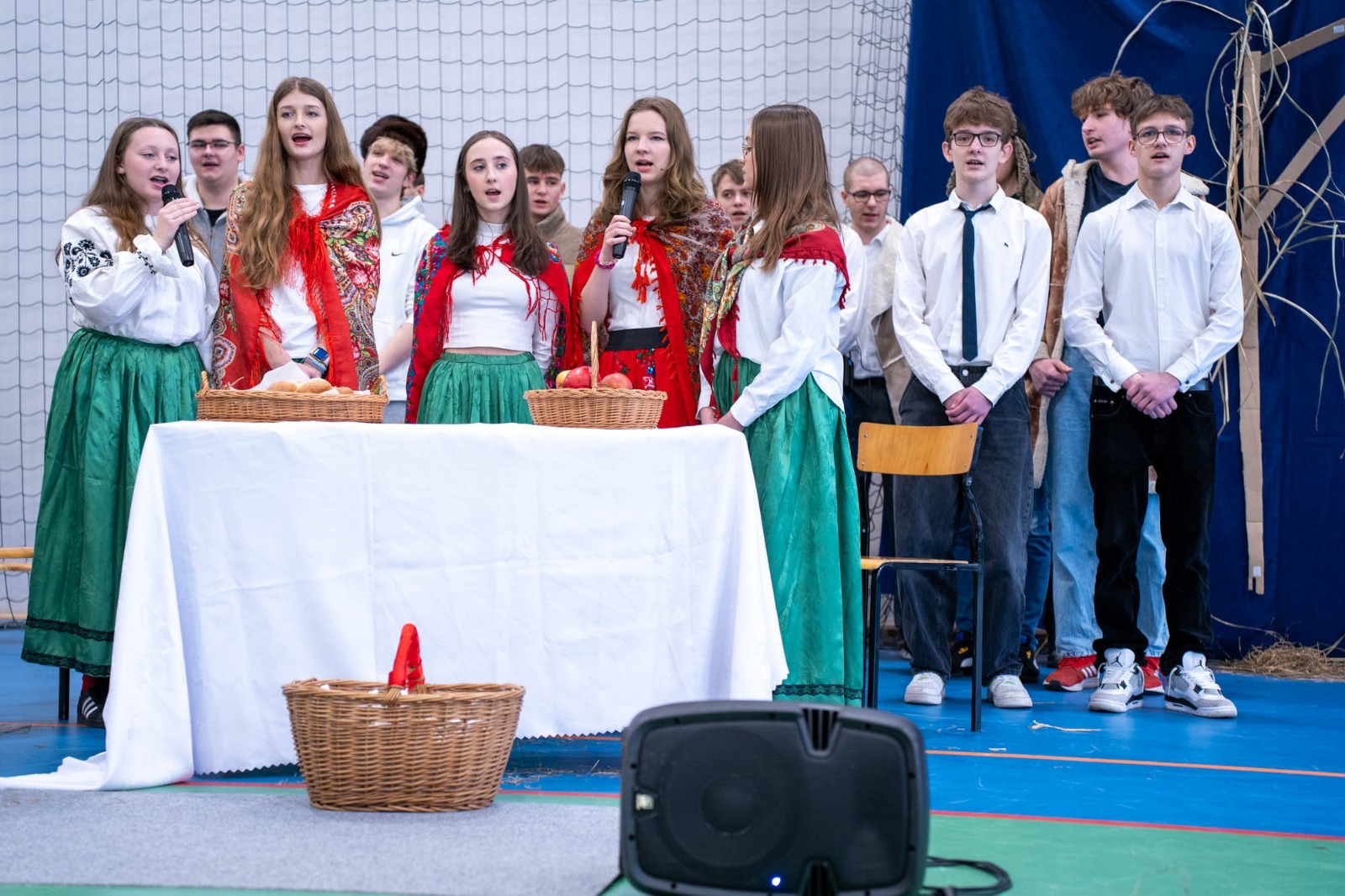 Uczniowie w trakcie przedstawienia: dziewczynki w strojach ludowych, a chłopcy ubrani na galowo stoją przy stole z białym obrusem. 