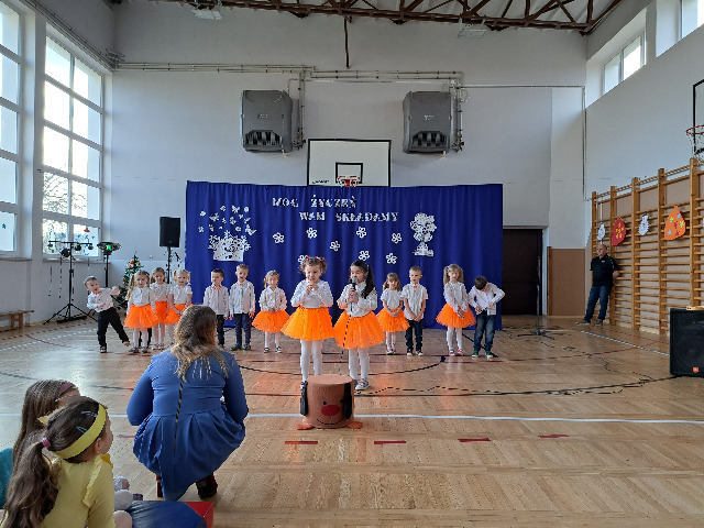 Występ przedszkolaków 4-5 latków na tle niebiesko białej dekoracji na dzień Babci i Dziadka.