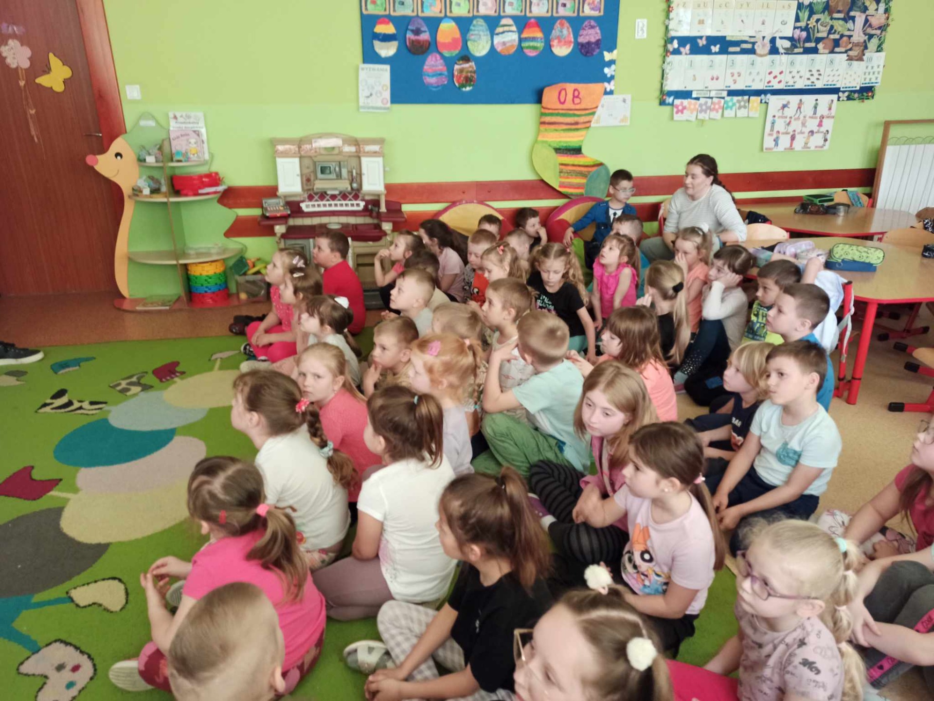 Przedszkolaki siedzą na kolorowym dywanie i słuchają opowieści gości.