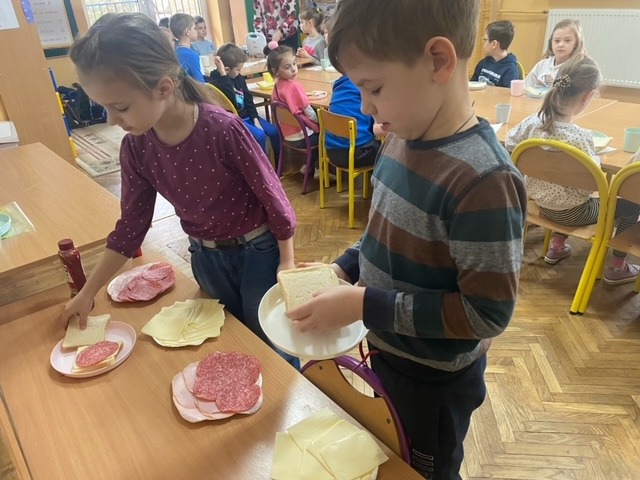 Uczeń i uczennica przekładają pieczywo tostowe ulubioną wędliną i żółtym serem. W tle uczniowie klasy pierwszej 
