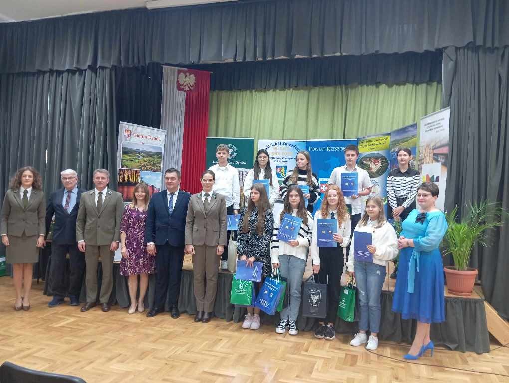 Na zdjęciu znajduje się szesnaście osób - organizatorzy i laureaci Wojewódzkiego Konkursu Ortograficznego "Leśne Dyktando". Za nimi stoją banery i flaga Polski.
