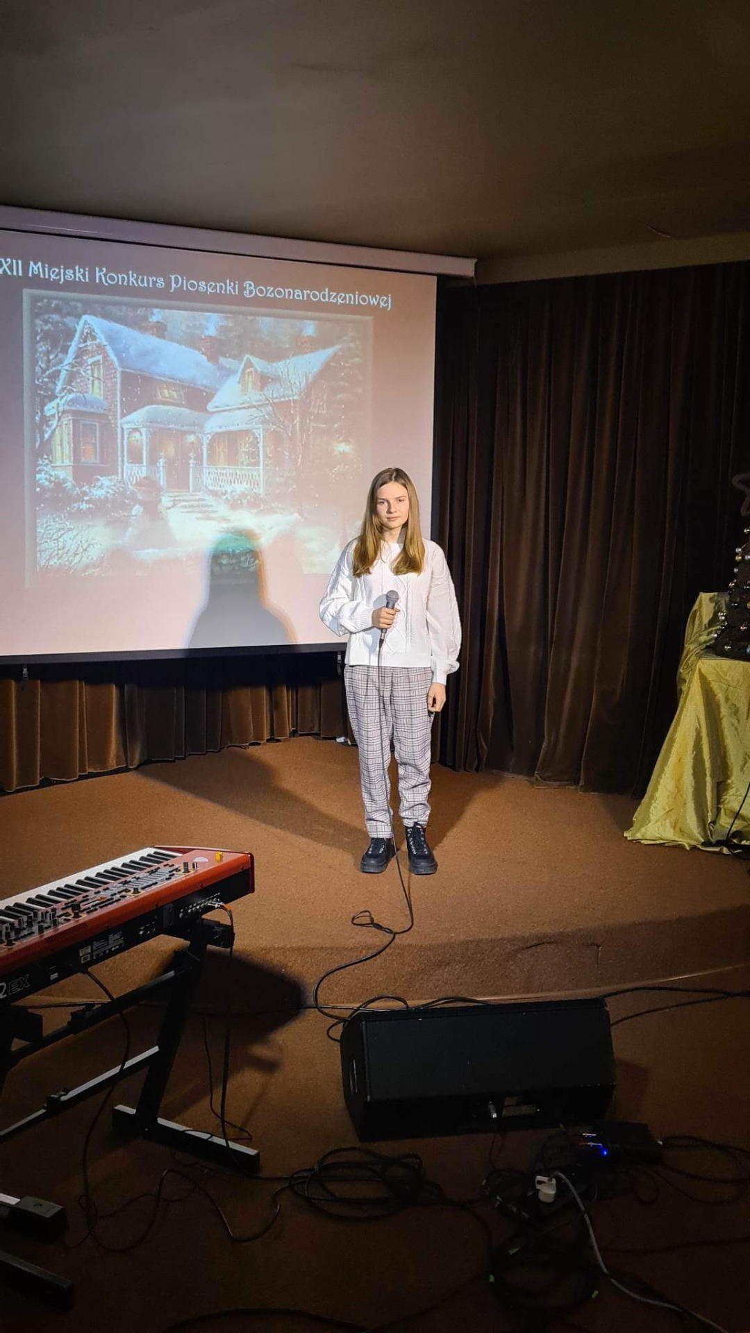 Dziewczyna w białym sweterku i spodniach w kratkę stoi na scenie. W ręku trzyma mikrofon. W tle widoczna dekoracja: prezentacja z motywami świątecznymi, choinka, pianino.