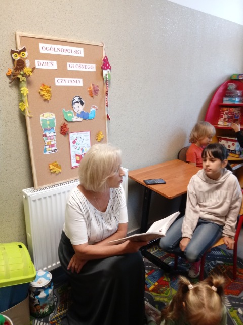 Nauczycielka siedzi i trzyma w prawej ręce otwartą książkę. Obok niej siedzi dziewczynka.