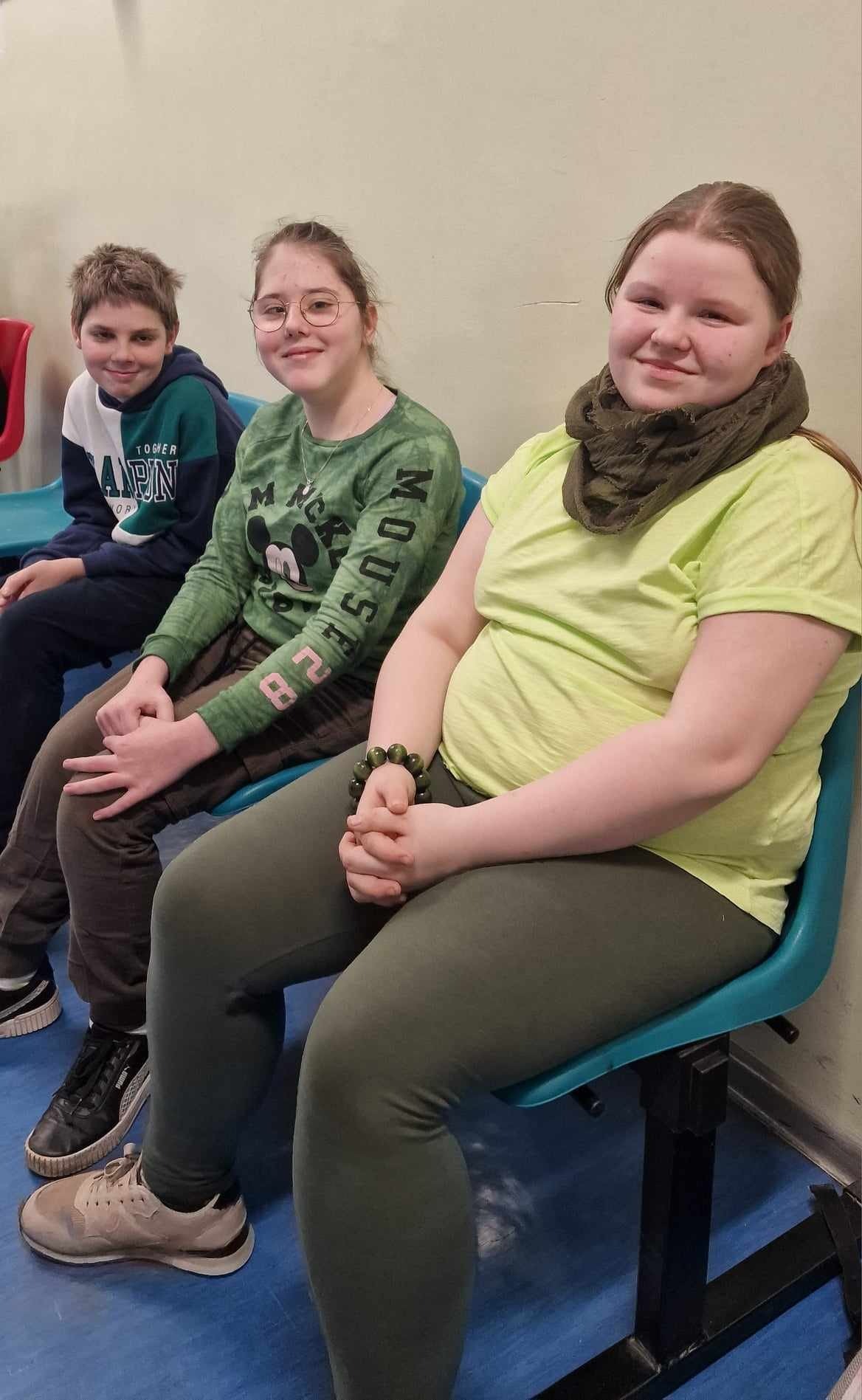 3 uczniów (chłopiec i 2 dziewczynki) siedzą na łąwkach uśmiechają się i pozują do zdjęciach, mają zielone elemnty stroju