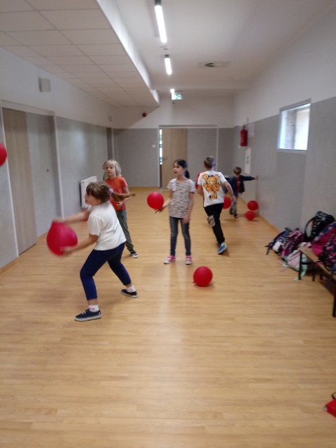 Grupka dzieci biega po korytarzu szkolnym, a pomiędzy nimi czerwone balony.