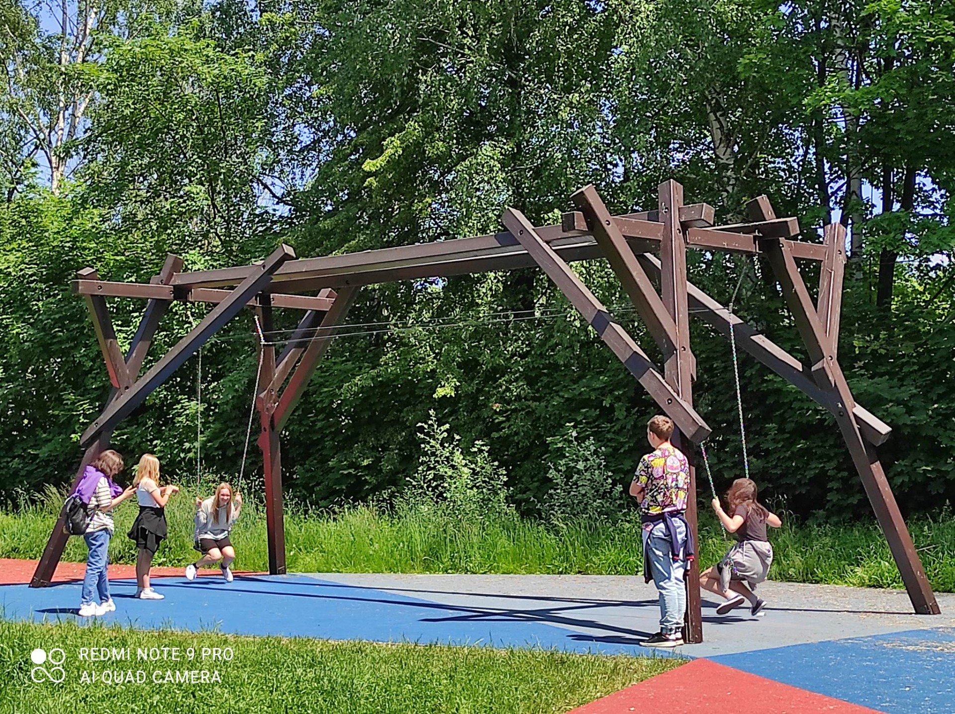 Zdjęcie przedstawia uczniów w Parku Doświadczeń. Dwie dziewczynki siedzą na huśtawkach. Po prawej stronie na kolorowym chodniku stoi chłopiec a po lewej dwie dziewczynki, które obserwują zabawę. W tle drzewa.